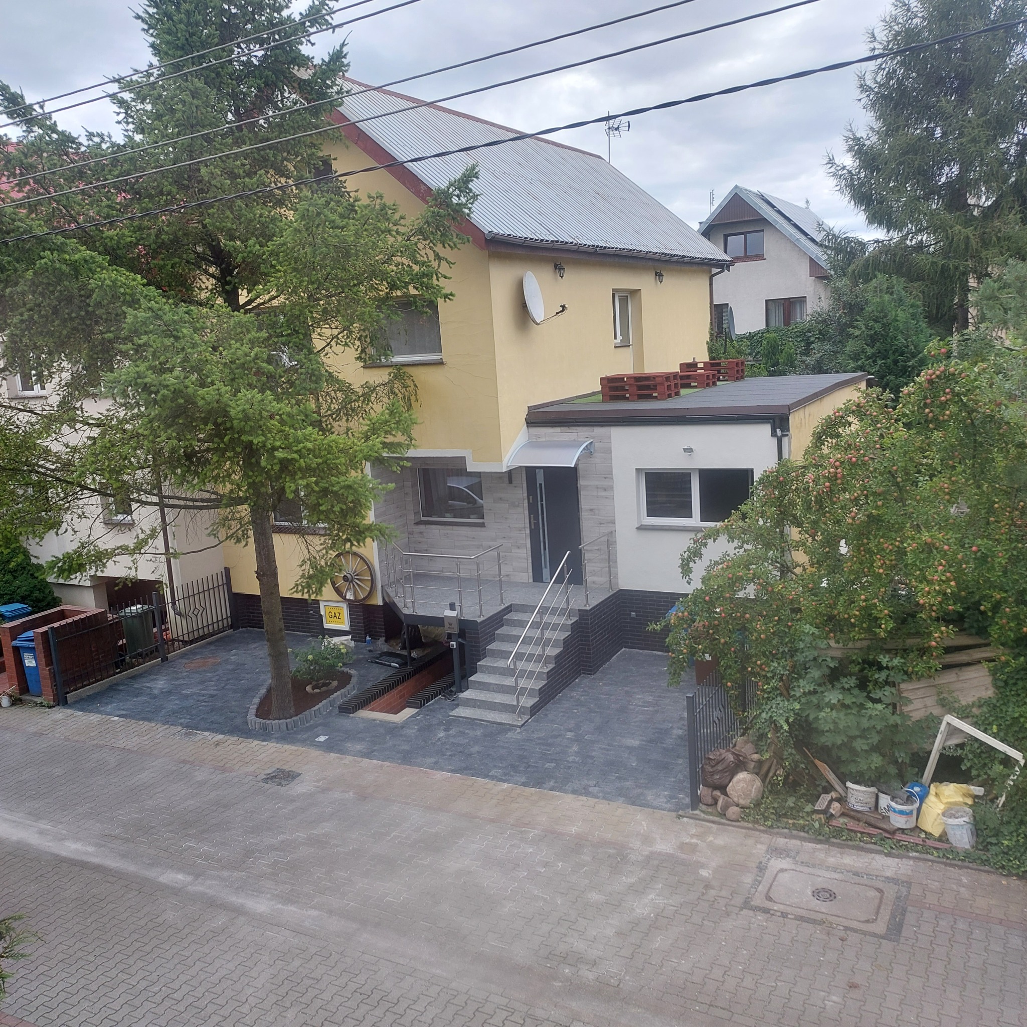 Żółty dom jednorodzinny z szarym dachem, nową elewacją z elementami dekoracyjnymi i podjazdem wyłożonym kostką brukową w dwóch odcieniach szarości, widok z góry.