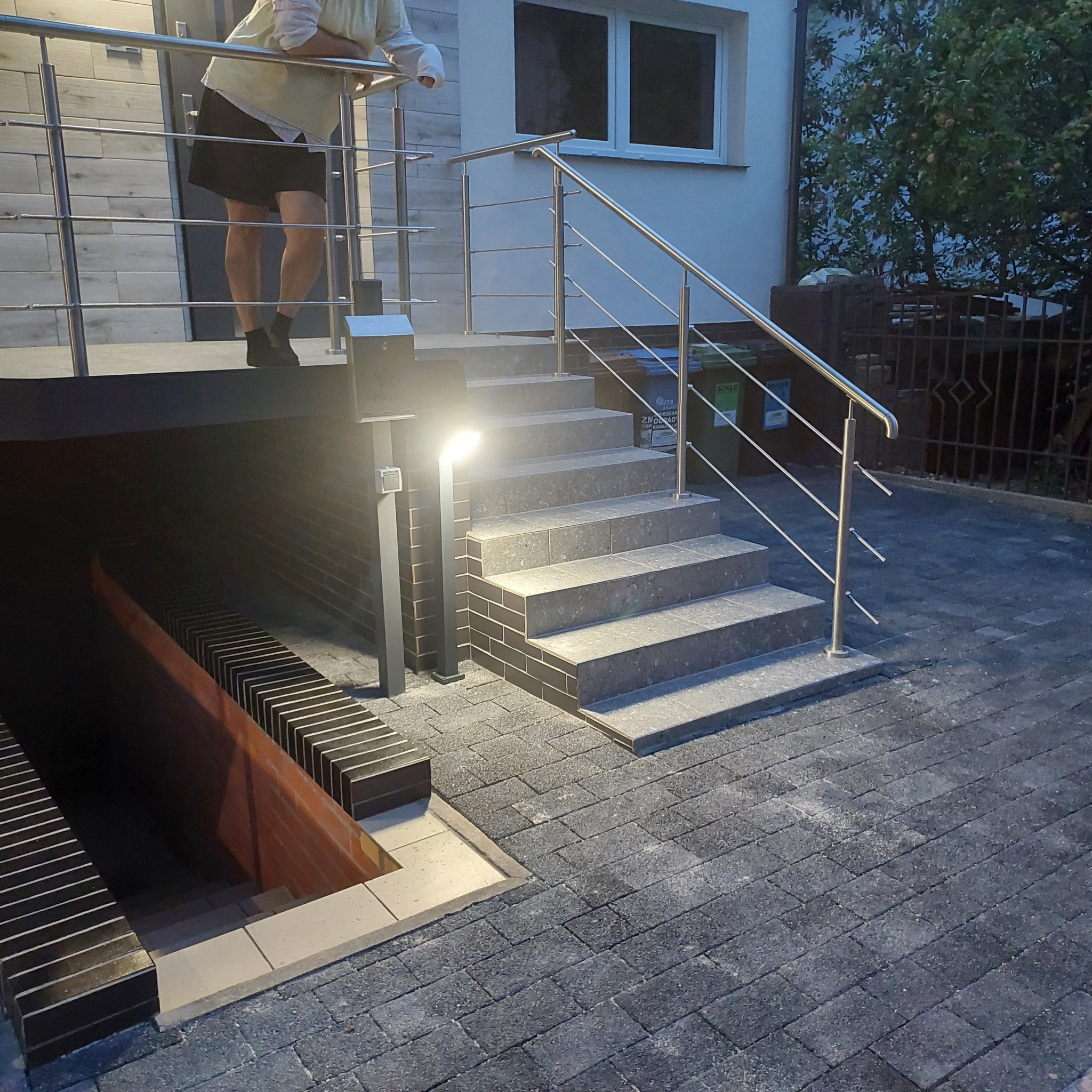 Zewnętrzne schody z szarej kostki brukowej prowadzące do piwnicy i na taras, z metalową balustradą i oświetleniem punktowym, widok na dom z oknami.