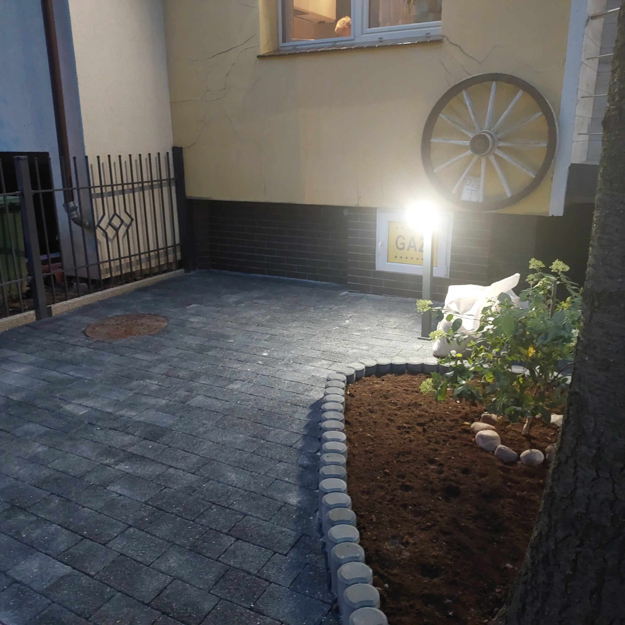 Ułożona szara kostka brukowa na podwórku z obrzeżem oddzielającym rabatę z krzewem, na ścianie budynku dekoracyjne koło od wozu.