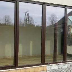 Zabudowa tarasu z brązowymi ramami okiennymi, widoczne odbicia drzew i słupów energetycznych w szybach, jasna elewacja budynku.