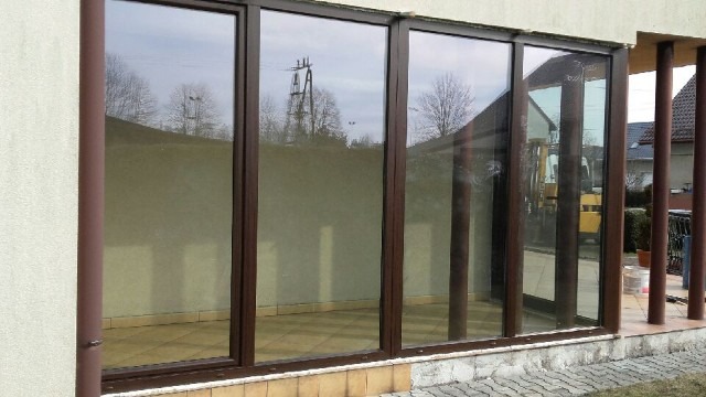 Zabudowa tarasu z brązowymi ramami okiennymi, widoczne odbicia drzew i słupów energetycznych w szybach, jasna elewacja budynku.