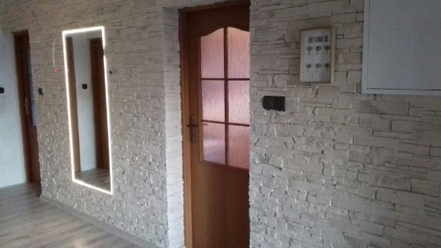 Wnętrze po remoncie z dekoracyjną ścianą wyłożoną jasnym kamieniem, drewniane drzwi z szybą, podświetlona wnęka w ścianie oraz drewniana podłoga.