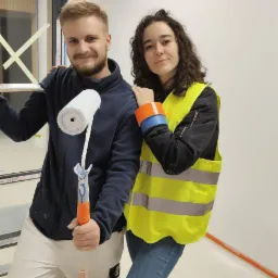 Uśmiechnięty malarz z wałkiem i kobieta w kamizelce odblaskowej, świeżo pomalowana biała ściana w tle, widoczne ślady farby na ubraniu malarza.