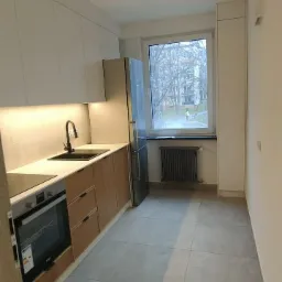 Nowoczesna kuchnia z drewnianymi frontami szafek, czarnym zlewem i baterią, szarą podłogą, oświetleniem punktowym i widokiem na park z okna.