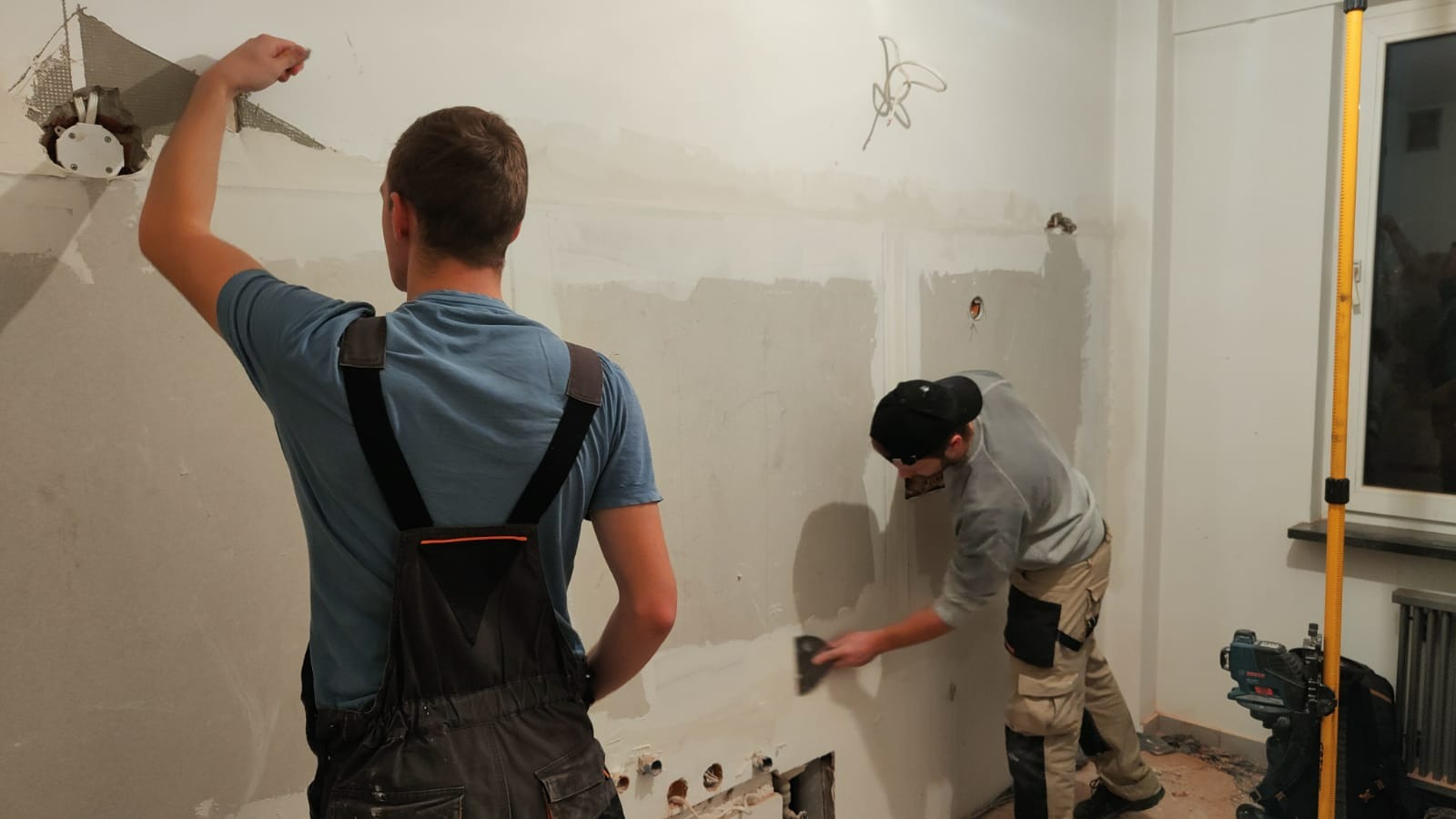 Dwóch mężczyzn w trakcie szpachlowania ściany w pomieszczeniu, widoczne niedoskonałości i otwory w ścianie, jeden z mężczyzn w kombinezonie i niebieskiej koszulce, drugi w czapce z daszkiem...