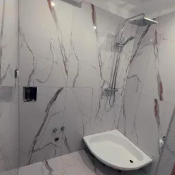 Nowoczesna łazienka z białymi płytkami imitującymi marmur z brązowymi żyłami, narożny brodzik, chromowana bateria prysznicowa z deszczownicą i otwór w ścianie z widoczną instalacją.