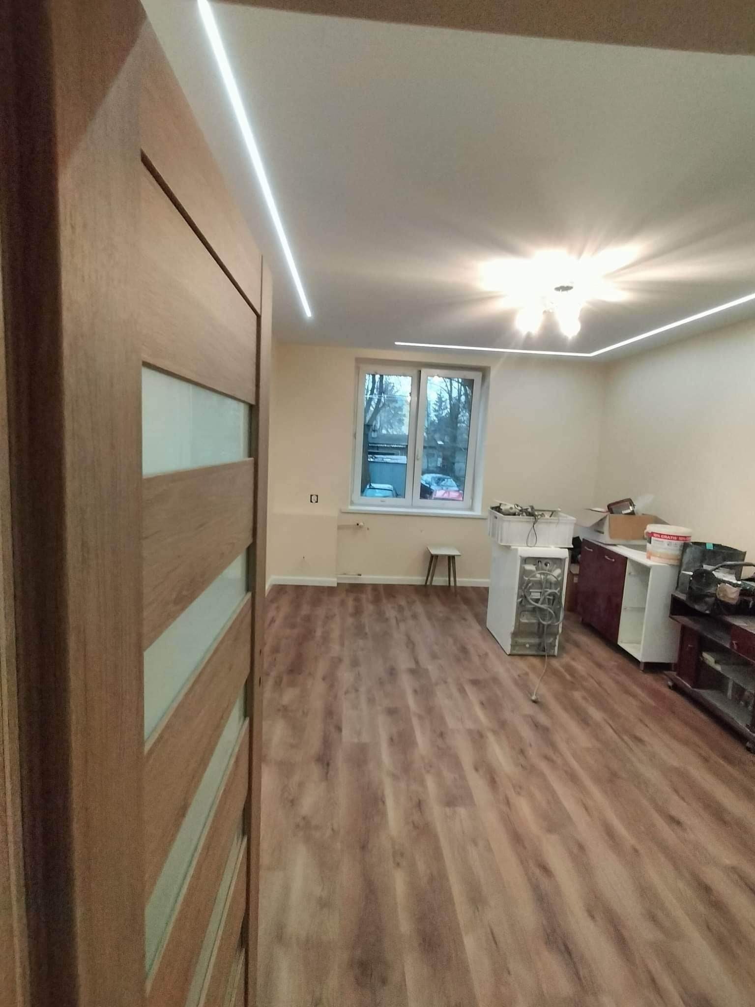 Wnętrze pokoju po remoncie z drewnianą podłogą, widoczne drzwi z przeszkleniami, listwy LED pod sufitem, okno i meble w tle.