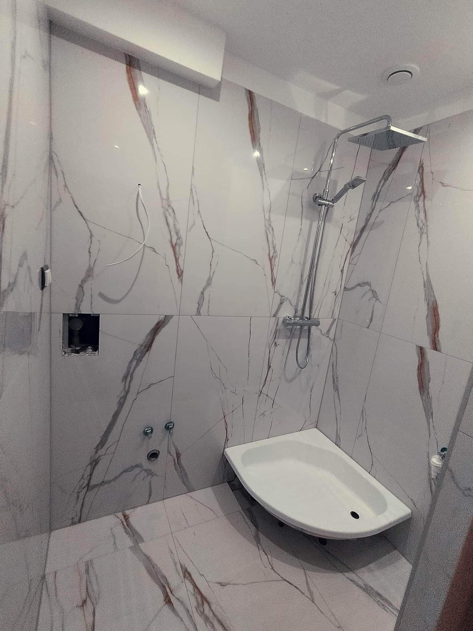 Nowoczesna łazienka z białymi płytkami imitującymi marmur z brązowymi żyłami, narożny brodzik, chromowana bateria prysznicowa z deszczownicą i otwór w ścianie z widoczną instalacją.