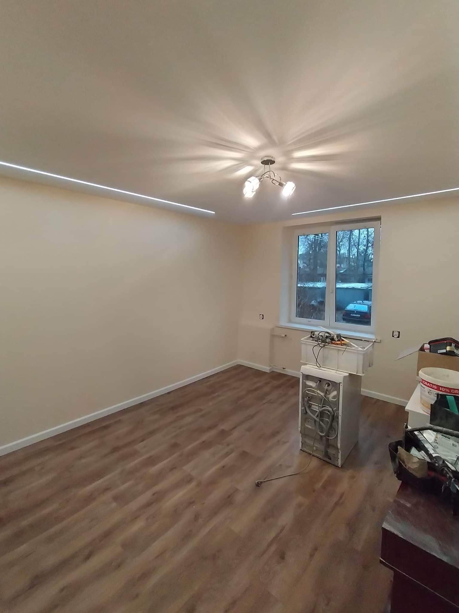 Wnętrze pokoju w trakcie remontu, z widocznym odnowionym parkietem, świeżo pomalowanymi ścianami, liniowym oświetleniem LED pod sufitem i przygotowanym do instalacji sprzętem AGD oraz narzędziami.