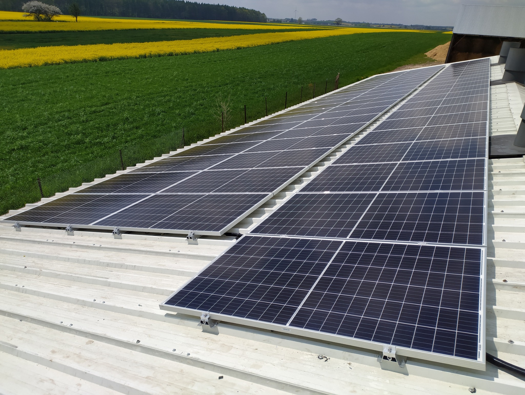 Rząd paneli słonecznych na blaszanym dachu z widokiem na pola uprawne w oddali, z uwzględnieniem metalowych uchwytów mocujących.