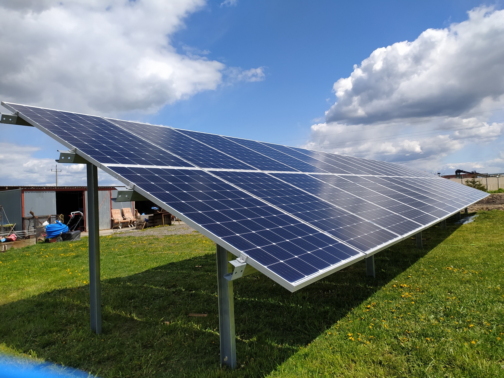 Rząd paneli słonecznych na metalowej konstrukcji, zainstalowany na zielonej trawie z widocznym budynkiem gospodarczym w tle i błękitnym niebem z chmurami.