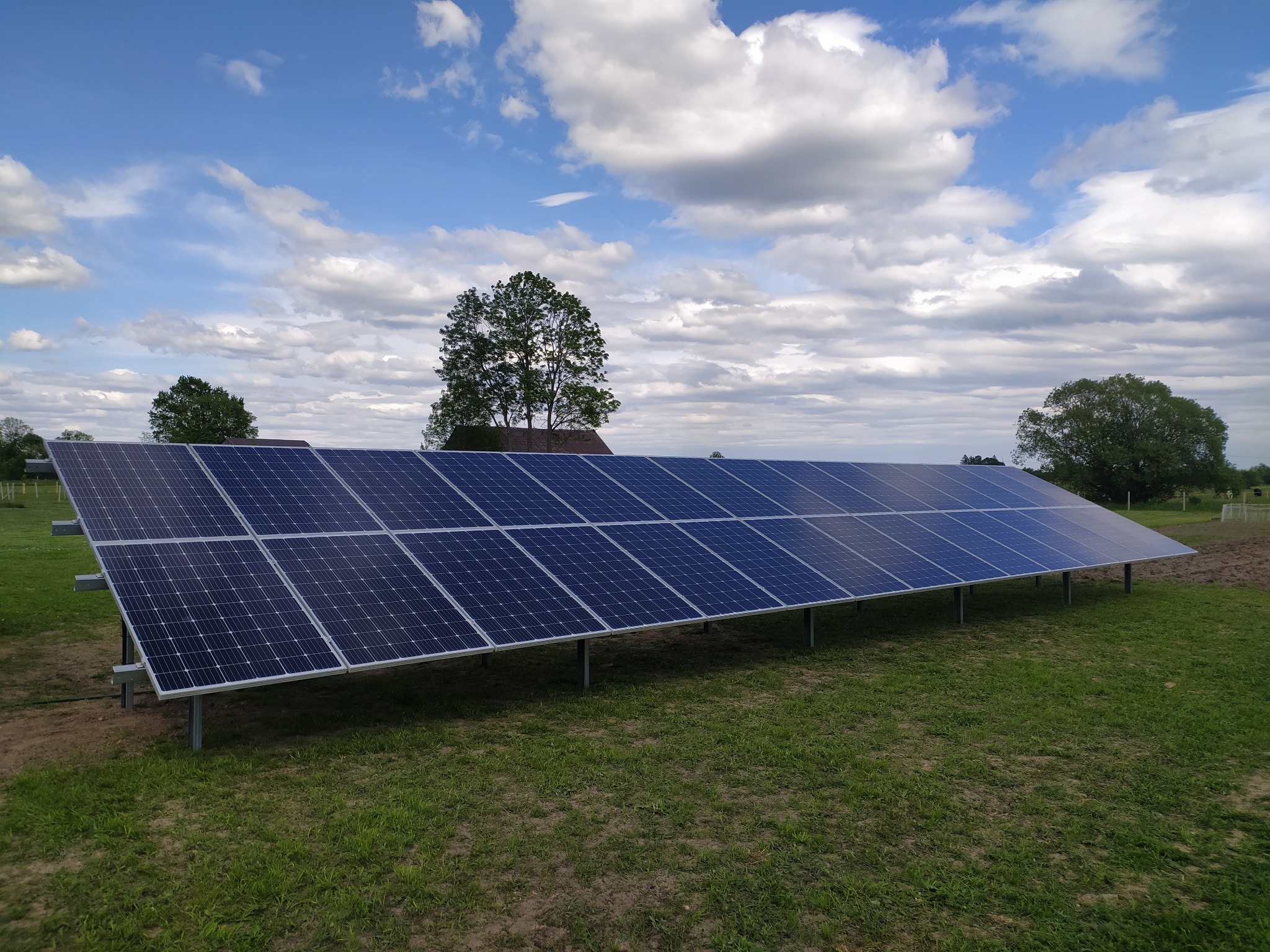 Rząd paneli słonecznych zamontowanych na metalowych wspornikach na trawiastym terenie, w tle drzewa i błękitne niebo z chmurami.