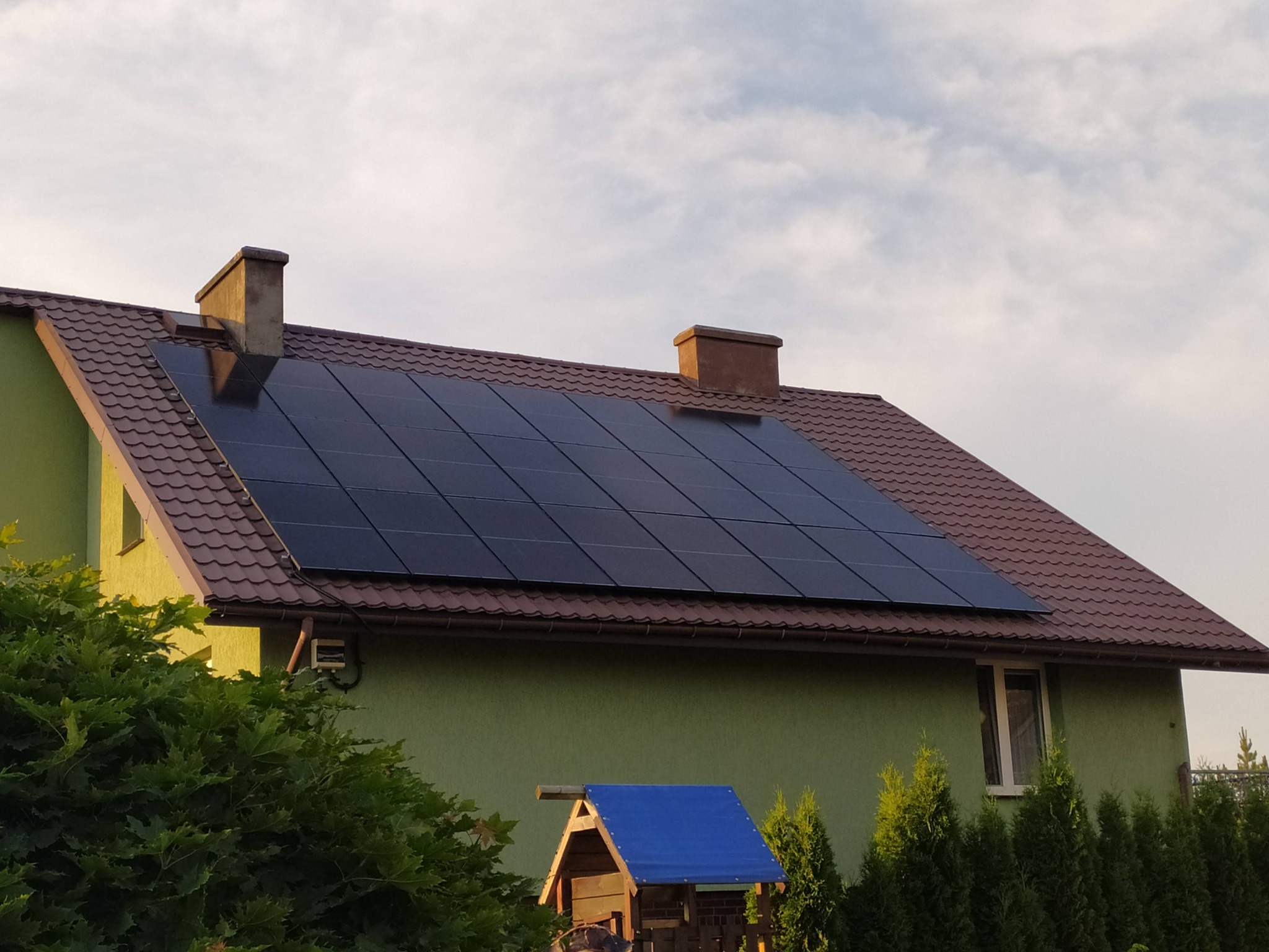 Panele słoneczne na brązowym dachu zielonego domu z dwoma kominami, widoczne z perspektywy ogrodu z małym, niebieskim domkiem zabaw.
