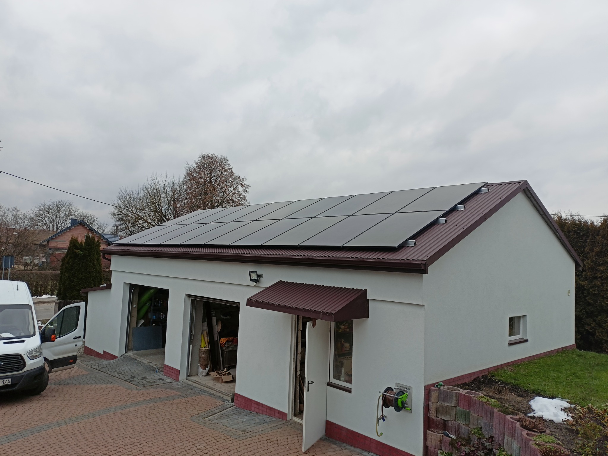 Panele słoneczne na dachu garażu z dwoma bramami, obok zaparkowany biały bus.