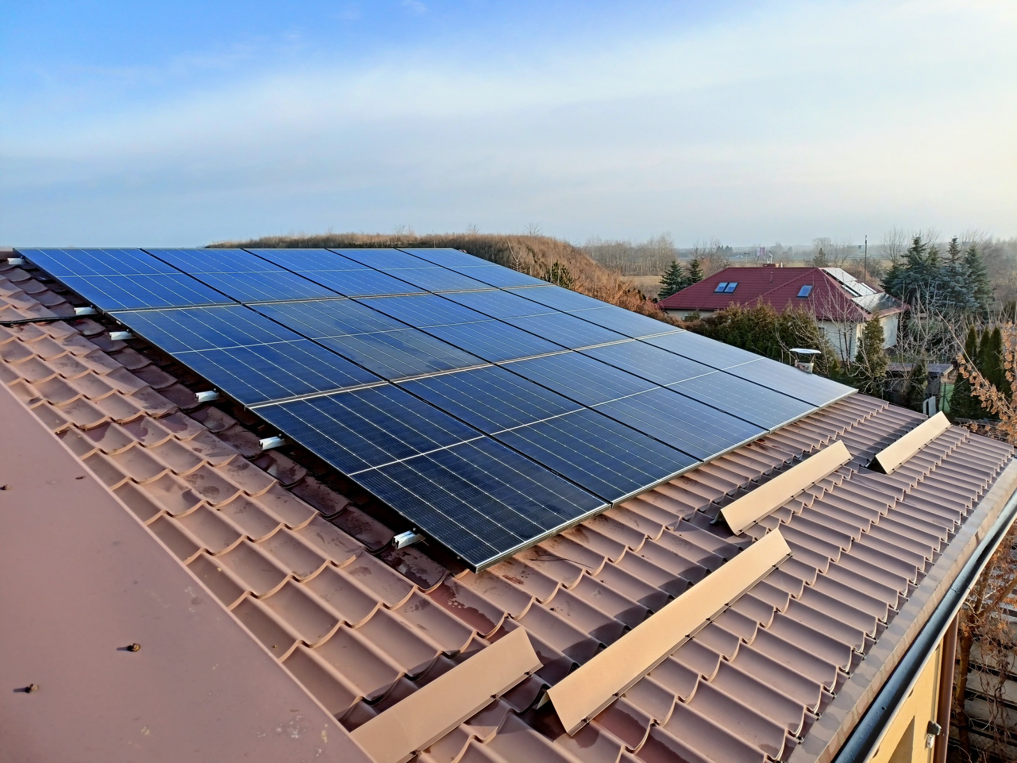 Panel słoneczny na brązowym dachu z dachówki ceramicznej, widok z góry na tle błękitnego nieba i zabudowań.