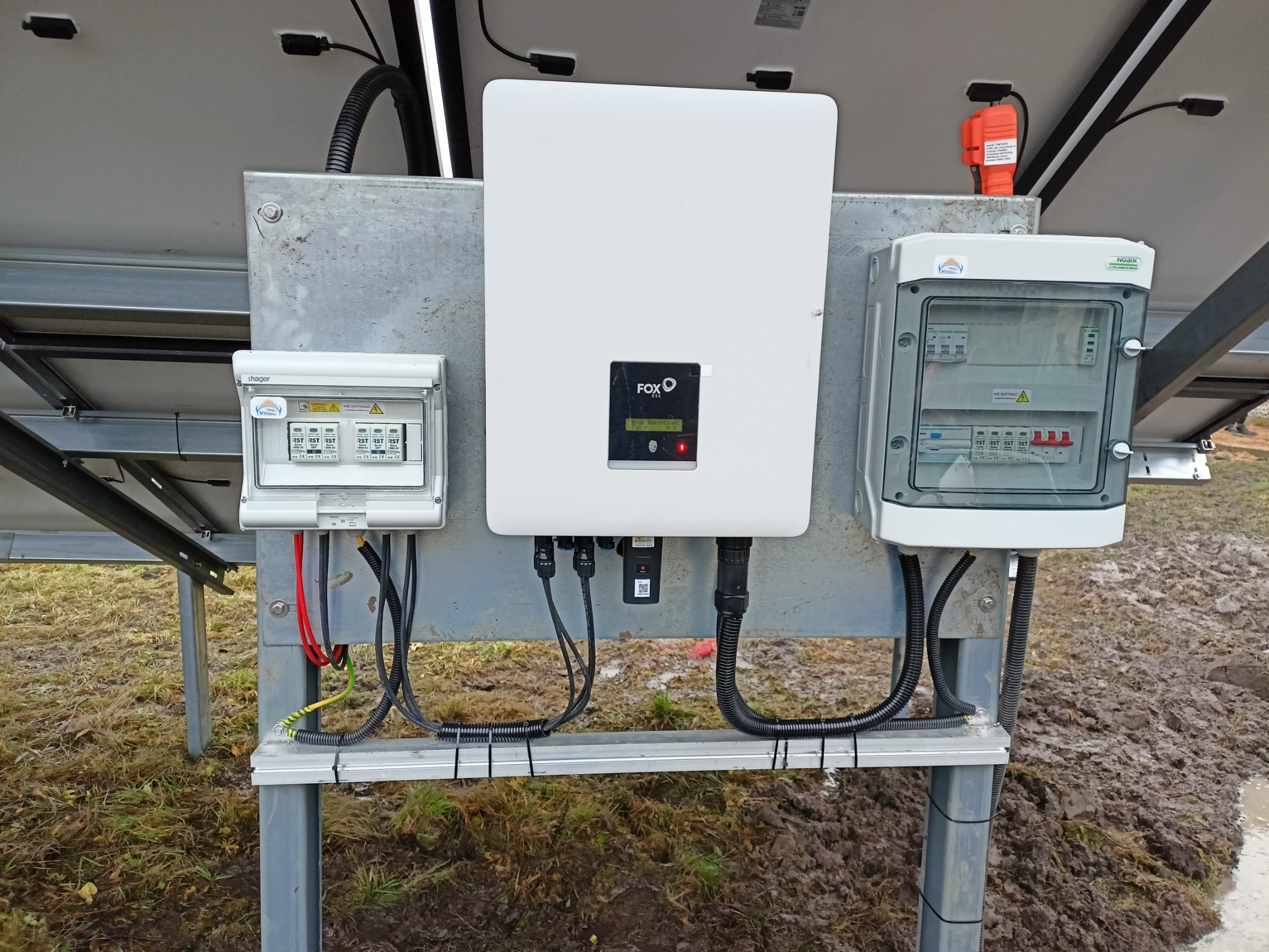 Instalacja paneli słonecznych z widocznym falownikiem FOX i dwoma skrzynkami zabezpieczeń na metalowej konstrukcji, na tle błotnistego terenu.