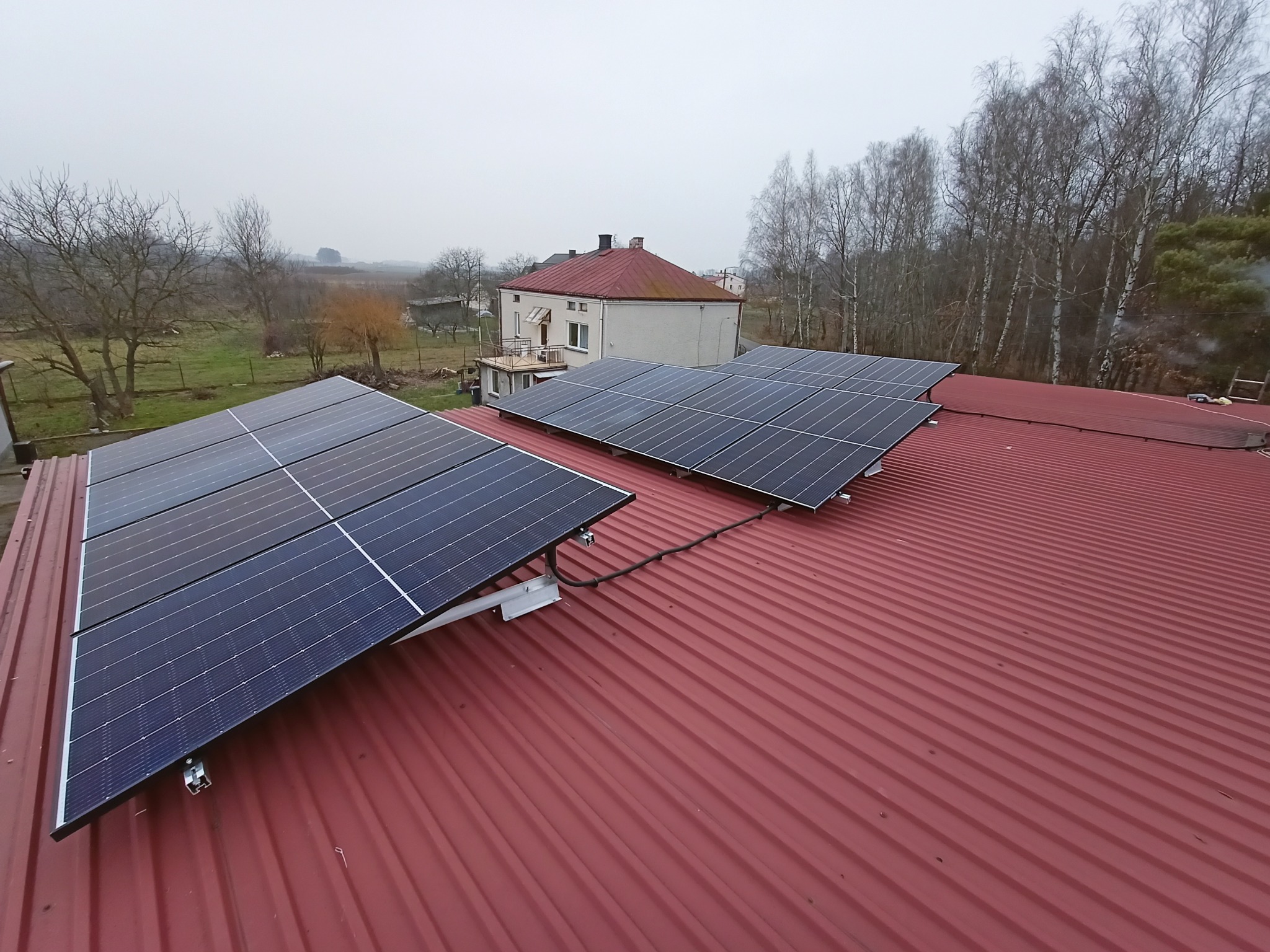Panele słoneczne zamontowane na czerwonym blaszanym dachu budynku jednorodzinnego w pochmurny dzień. Widoczne mocowania paneli i okablowanie. W tle dom i drzewa bez liści.