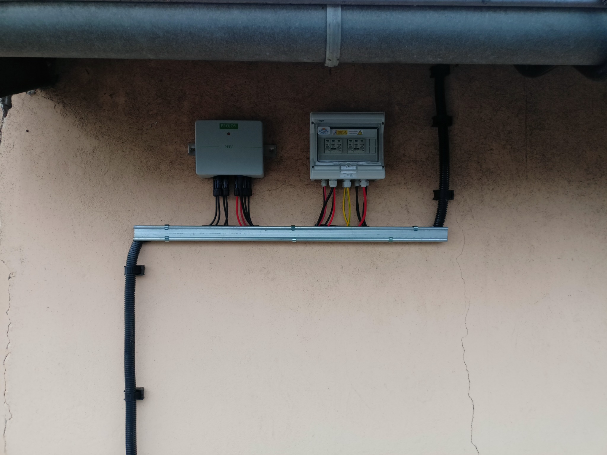 Zamontowana na zewnątrz skrzynka elektryczna z przewodami, podłączona do falownika, przymocowana do ściany budynku za pomocą szyny montażowej.