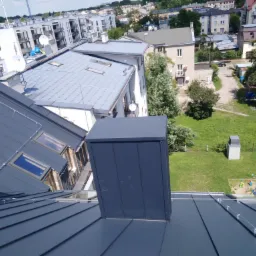 Grafitowy dach z blachy na rąbek stojący, z widocznym kominem i zabezpieczeniem przeciwśniegowym, na tle zabudowy miejskiej i zielonego ogrodu.