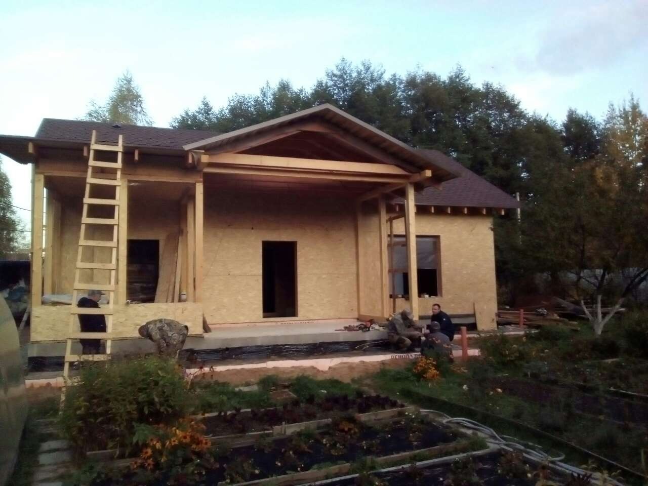 Budowa parterowego domu z drewnianą konstrukcją, widoczne fragmenty dachu z brązową dachówką, drabina oparta o ścianę, kilku robotników przy pracy na zewnątrz.