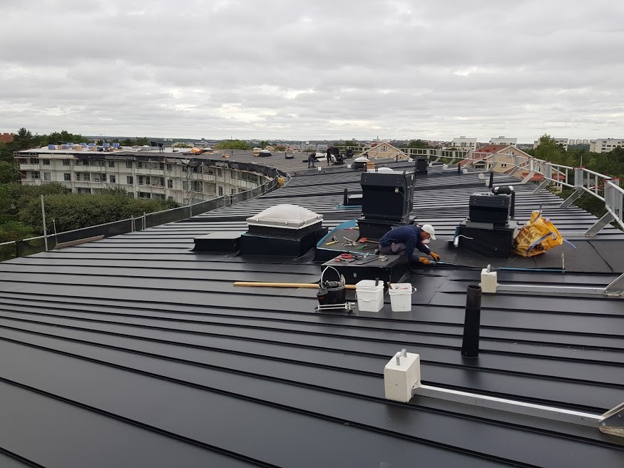 Prace na dachu z blachy na dużej powierzchni, widoczny dekarz w białej czapce przy pracy, narzędzia, wentylatory dachowe, świetlik i budynki w tle.