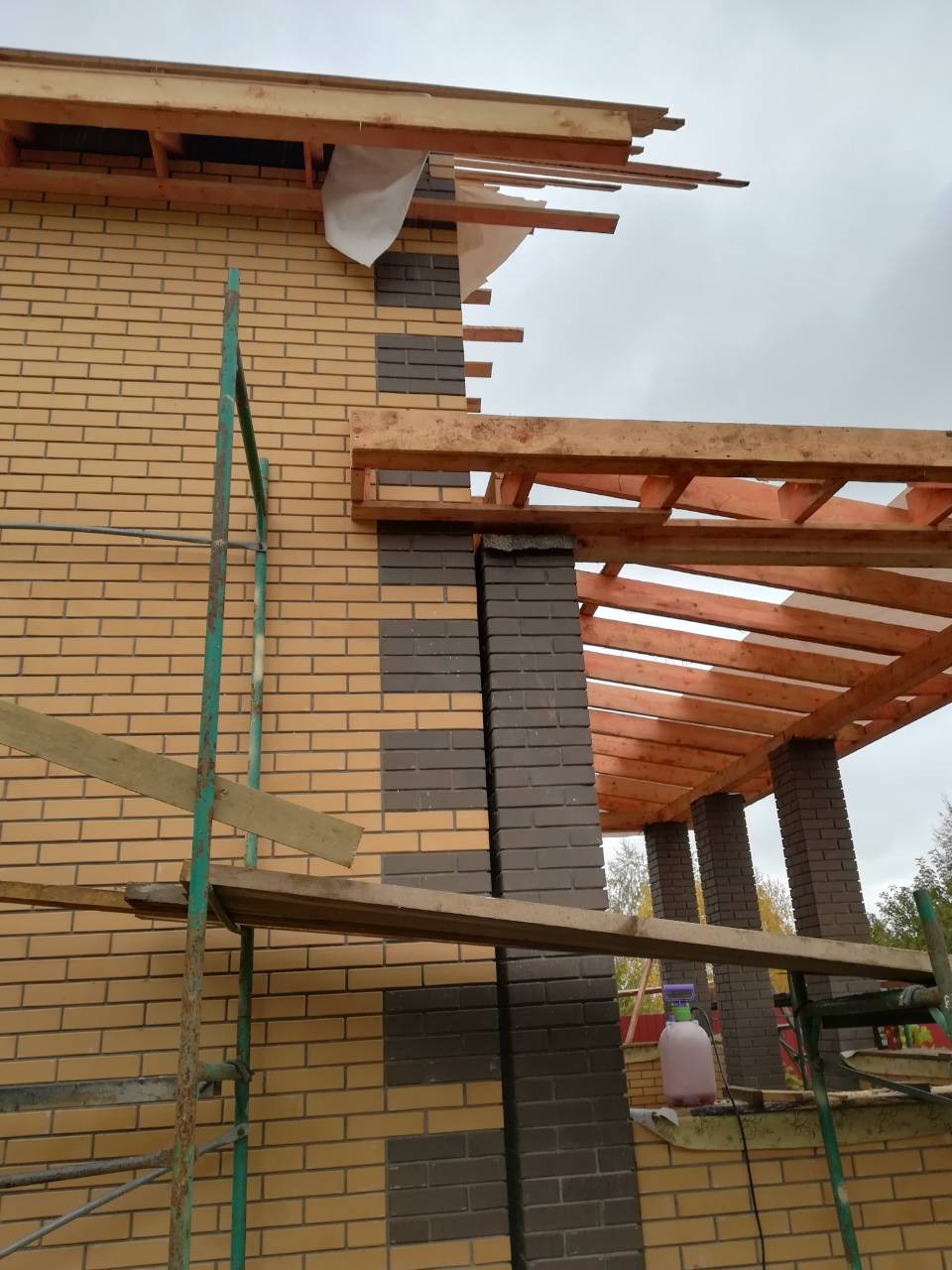 Budowa domu z cegły klinkierowej z drewnianą konstrukcją dachu w trakcie montażu, widoczne rusztowanie i narzędzia na tle pochmurnego nieba.