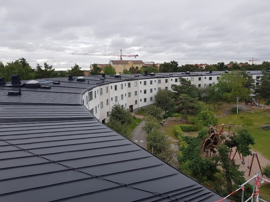 Czarny, blaszany dach wielorodzinnego budynku z kopułami wentylacyjnymi, widok z góry na park z placem zabaw i budynki w tle, pochmurne niebo.