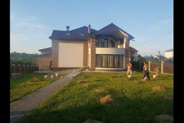 Nowoczesny dom jednorodzinny z brązową elewacją i ciemnym dachem, z trzema pracownikami przygotowującymi się do malowania elewacji na trawniku przed domem.