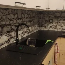 Szklany panel ścienny w kuchni z motywem kamiennej ściany, zamontowany nad czarnym blatem i zlewozmywakiem z baterią.