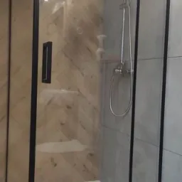 Nowoczesna kabina prysznicowa z czarnymi profilami i szklanymi ścianami, zamontowana w łazience z płytkami imitującymi drewno i szare płytki.