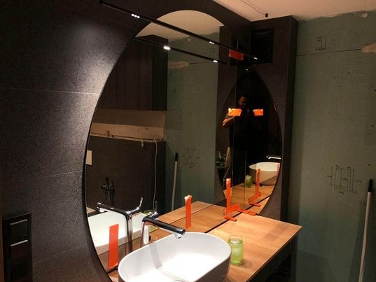 Owalne lustro w czarnej ramie z oświetleniem LED, odbijające fragment łazienki z umywalką i osobą robiącą zdjęcie telefonem, widoczne pomiary na ścianie.