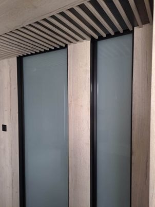 Matowe szklane panele z czarnymi ramami, wkomponowane w drewniane wykończenie ściany i sufitu z lamelami.