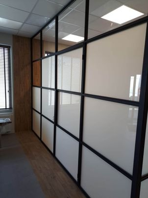 Przeszklona ściana działowa z czarnymi profilami w biurze, częściowo matowe szkło, widoczne rolety okienne.