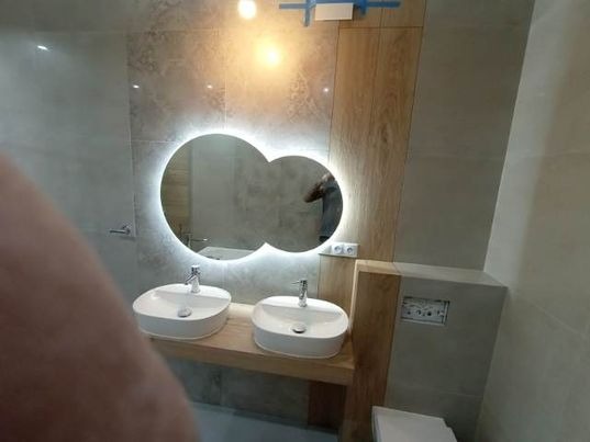 Nowoczesna łazienka z dwoma umywalkami nablatowymi o zaokrąglonym kształcie, podświetlanym lustrem w kształcie dwóch połączonych kół i drewnianym blatem oraz okładziną ściany.