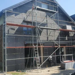 Budynek w trakcie prac termoizolacyjnych, widoczne rusztowanie, szare płyty izolacyjne i fragment ściany z cegły