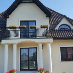 Wykończona elewacja domu jednorodzinnego w jasnym odcieniu z ciemną dachówką, balkonem z czarną balustradą i licznymi donicami z czerwonymi kwiatami.