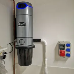 Szary odkurzacz centralny marki BEAM zamontowany na ścianie z widoczną instalacją rur miedzianych i gniazdem elektrycznym przemysłowym. Górna część urządzenia podświetlona na niebiesko.