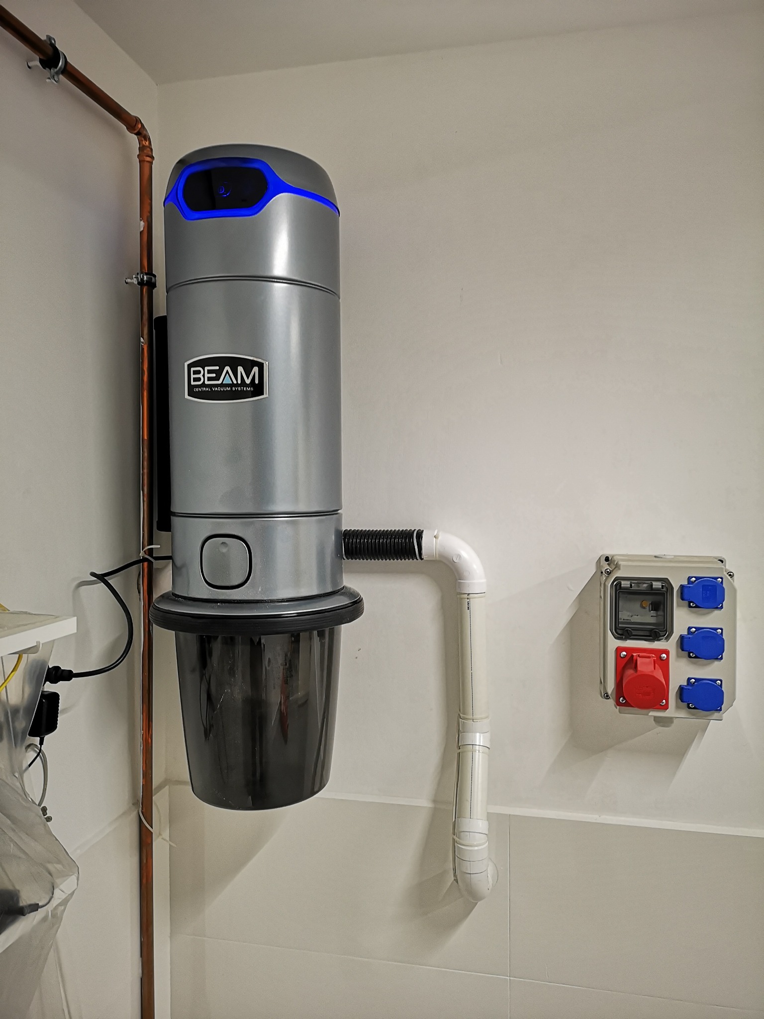 Szary odkurzacz centralny marki BEAM zamontowany na ścianie z widoczną instalacją rur miedzianych i gniazdem elektrycznym przemysłowym. Górna część urządzenia podświetlona na niebiesko.