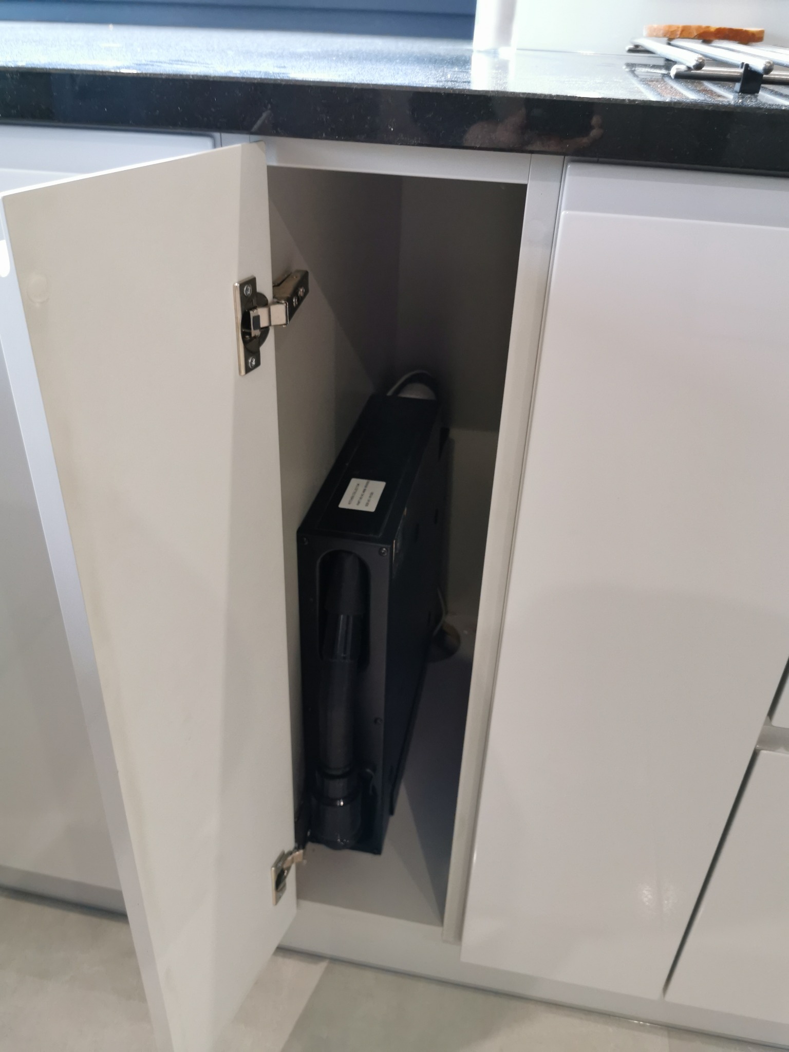 Czarny moduł centralnego odkurzacza, umieszczony w szafce pod blatem kuchennym z otwartymi drzwiczkami.