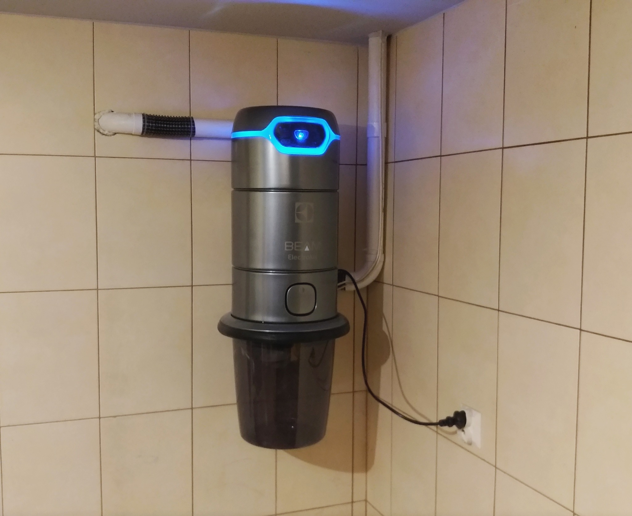 Zamontowany na kafelkowej ścianie szary odkurzacz centralny Beam Electrolux z podłączonym wężem i niebieskim podświetleniem.