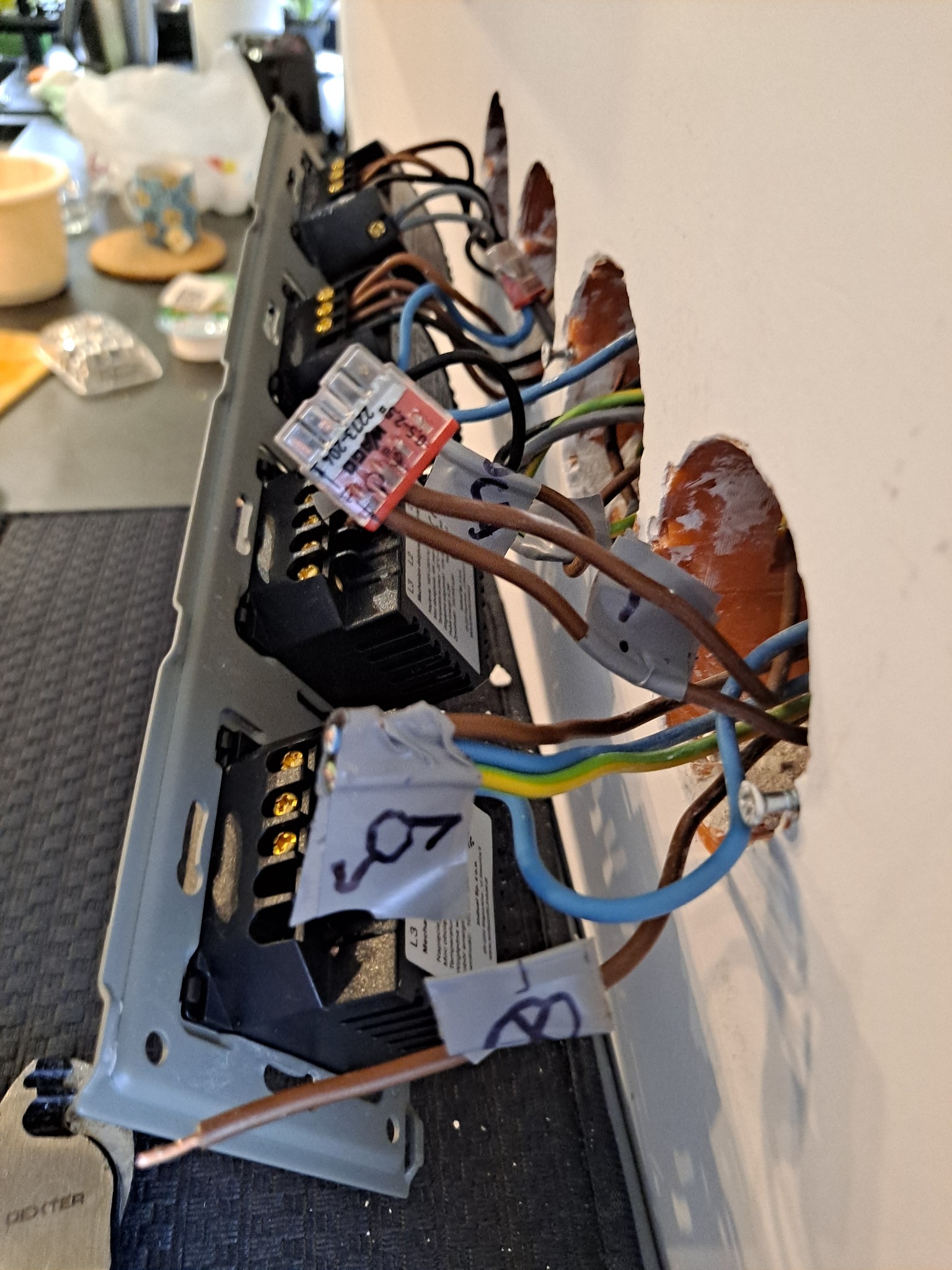 Przewody elektryczne w ścianie, podłączone do gniazd w metalowej ramce, zabezpieczone taśmą izolacyjną i złączkami WAGO, widoczne otwory w ścianie i wystające kable.