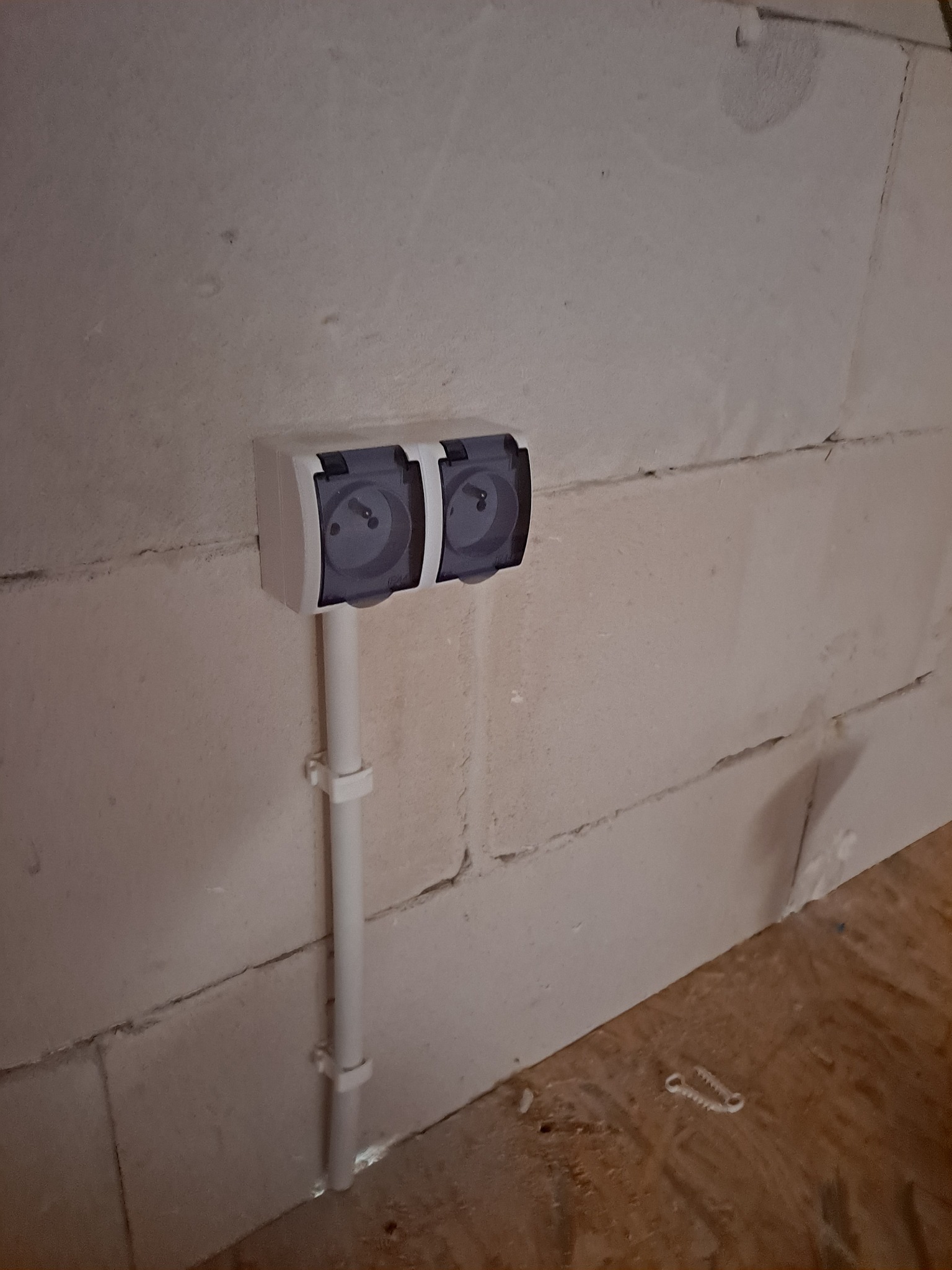 Podwójne gniazdko elektryczne natynkowe z klapkami, zamontowane na ścianie z bloczków betonowych. Rurka instalacyjna pionowo w dół, przymocowana uchwytami. Na podłodze leżą dwa plastikowe kołki...