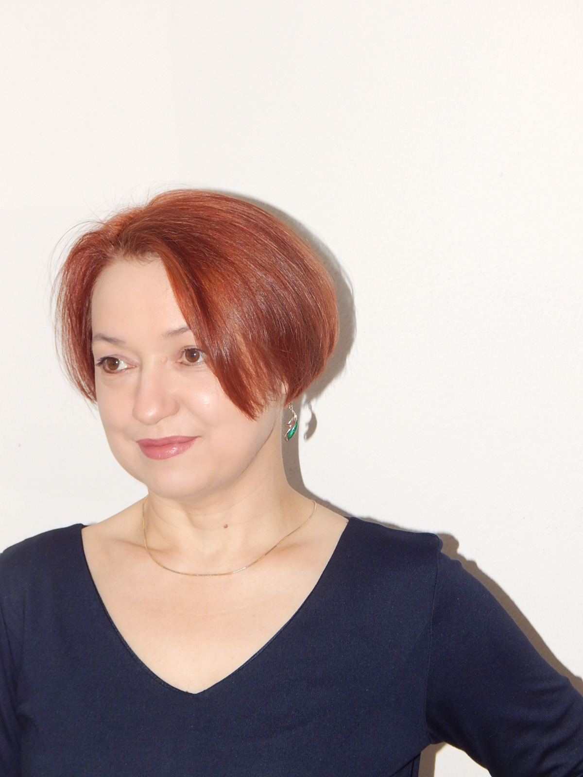 Portret kobiety z rudymi włosami, złotym łańcuszkiem i zielonymi kolczykami na tle białej ściany.