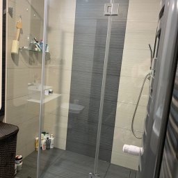 Vetus Art - Szklana kabina prysznicowa z chromowanymi okuciami, widok na półkę z kosmetykami i grzejnik drabinkowy w łazience wykończonej płytkami w odcieniach szarości i beżu.