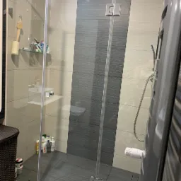 Szklana kabina prysznicowa z chromowanymi okuciami, widok na półkę z kosmetykami i grzejnik drabinkowy w łazience wykończonej płytkami w odcieniach szarości i beżu.