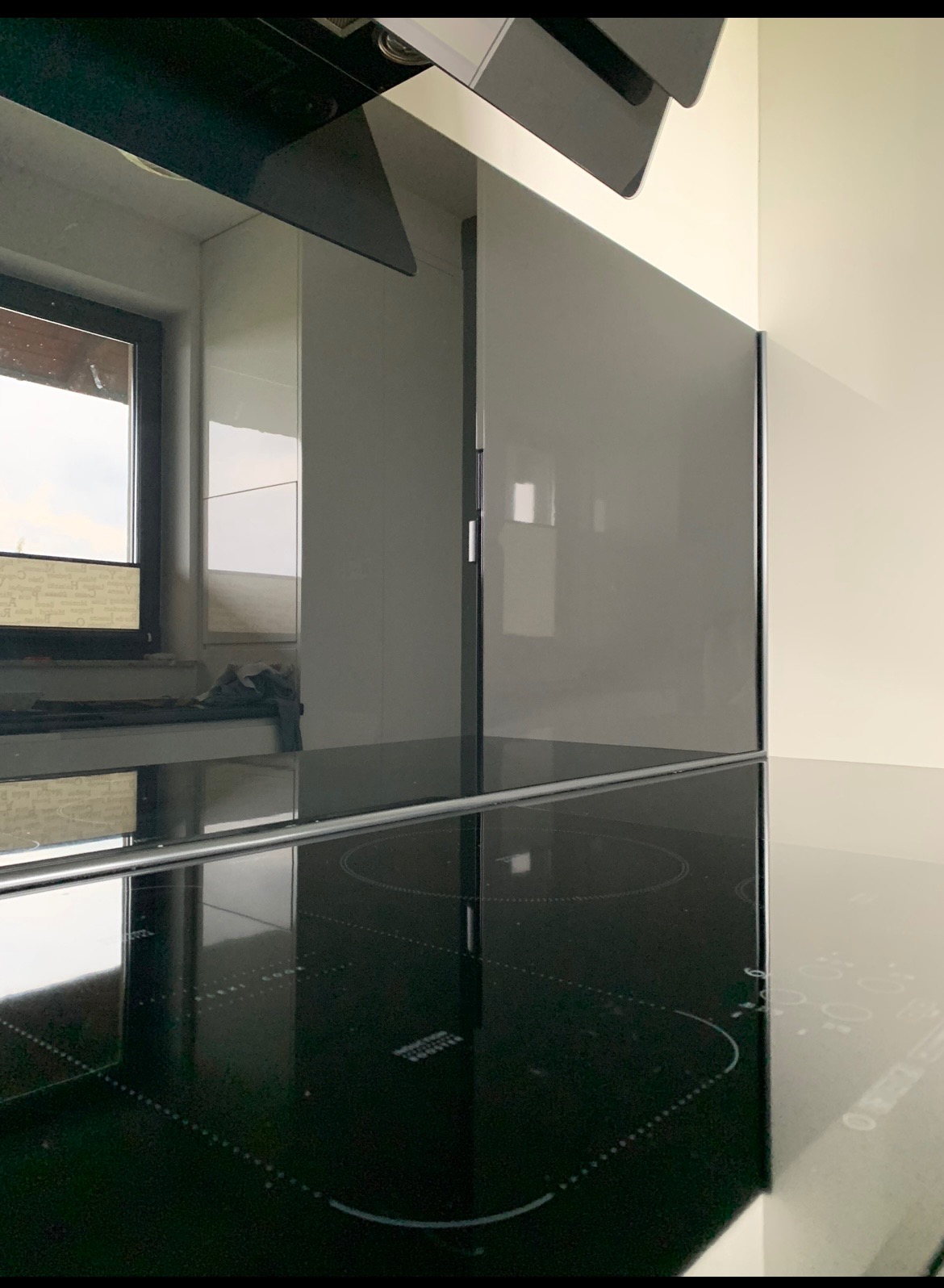 Błyszcząca, czarna płyta indukcyjna z widocznymi okręgami grzewczymi, odbijająca fragment okna i szarych szafek kuchennych, z okapem widocznym u góry.