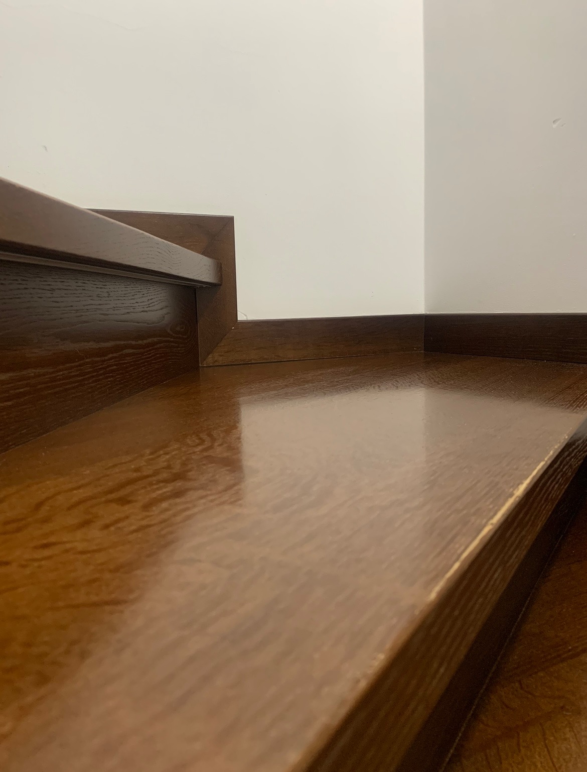 Błyszczące, lakierowane drewniane schody w kolorze ciemnego dębu, widok z dołu, przy białej ścianie z ciemną listwą przypodłogową.