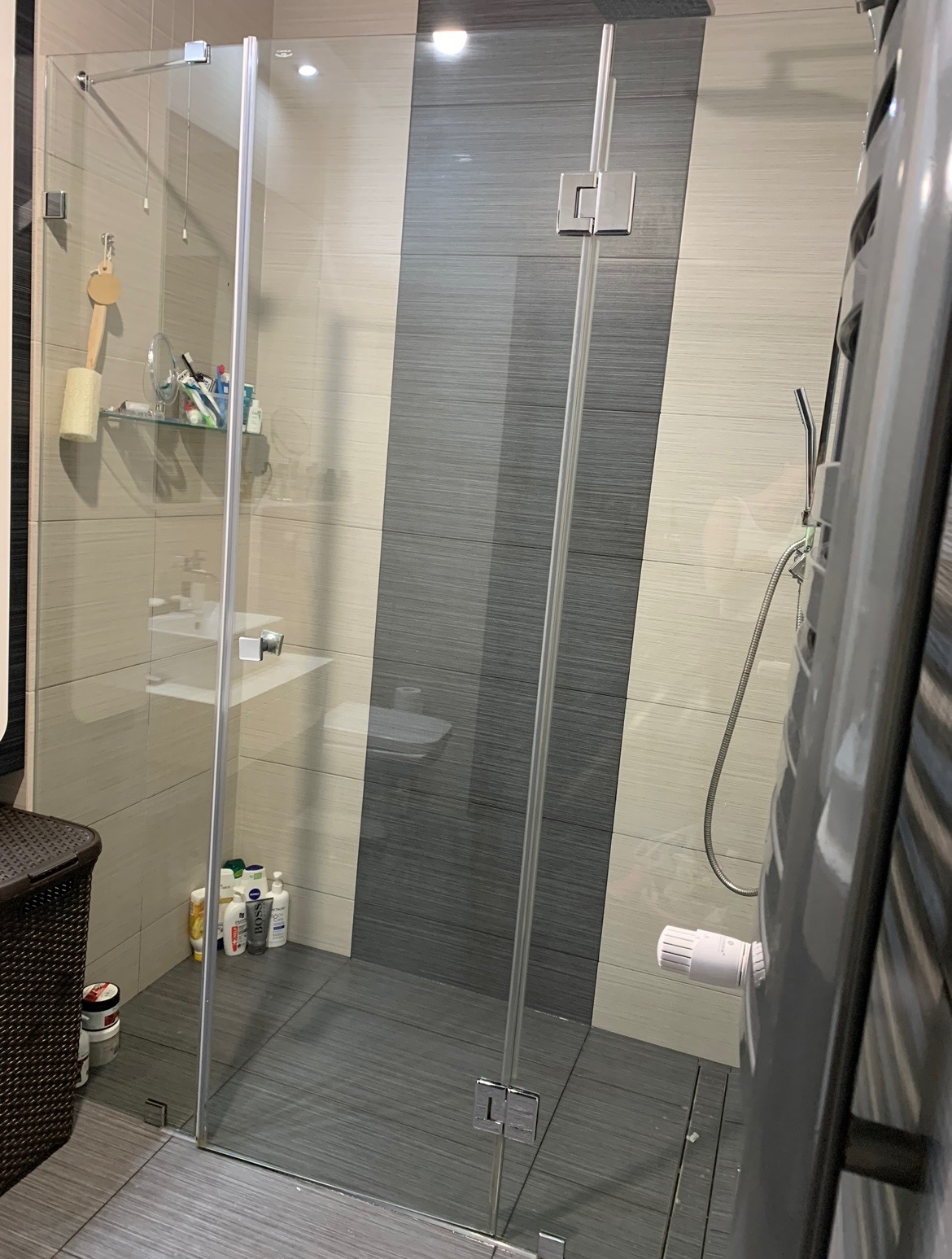 Szklana kabina prysznicowa z chromowanymi okuciami, widok na półkę z kosmetykami i grzejnik drabinkowy w łazience wykończonej płytkami w odcieniach szarości i beżu.