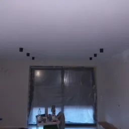 Sufit z zamontowanymi czarnymi reflektorami punktowymi, okno zabezpieczone folią malarską, ślady po remoncie na ścianach.