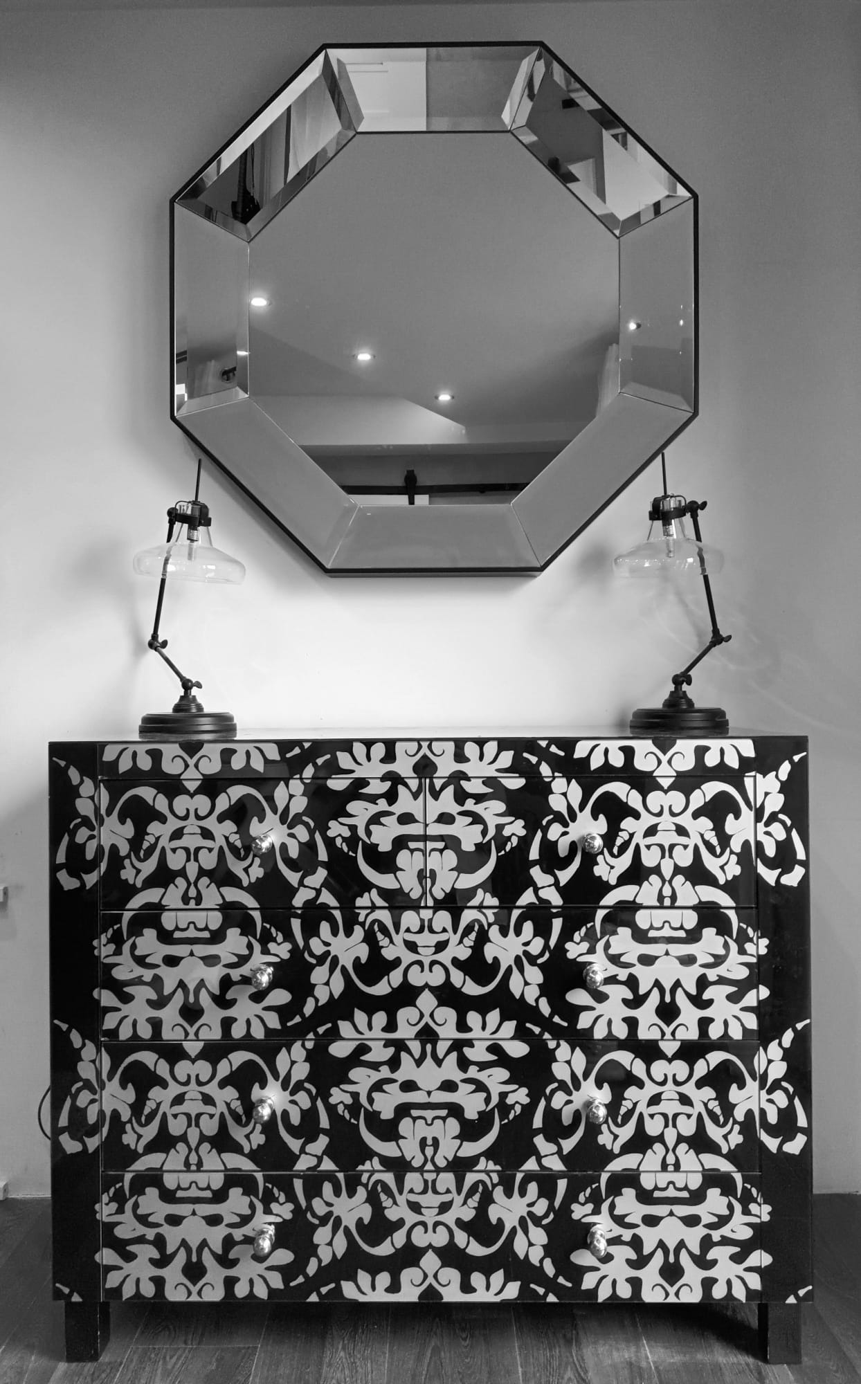 Czarno-biała komoda z ornamentem, zwieńczona ośmiokątnym lustrem i lampkami nocnymi ze szklanymi kloszami.